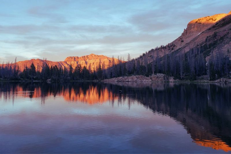 View across a lake with pine trees on the other side, with Uinta mountain peaks lit up at sunset.