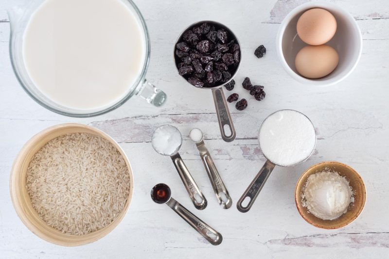 Ingredients for Instant Pot dairy free rice pudding including, almond milk, rice, vanilla, coconut oil, salt, sugar, corn starch, eggs, and dried cherries.