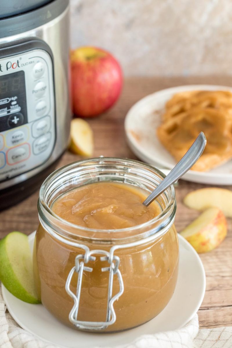 A glass jar filled with apple butter and placed in front of an Instant Pot, with waffles in the background.