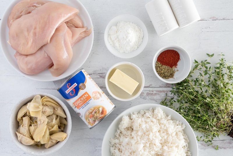 an overhead shot of all of the ingredients required to make artichoke chicken, including chicken breasts, artichoke hearts, chicken broth, butter, corn starch, salt and pepper, spices, thyme, and cooked rice for serving