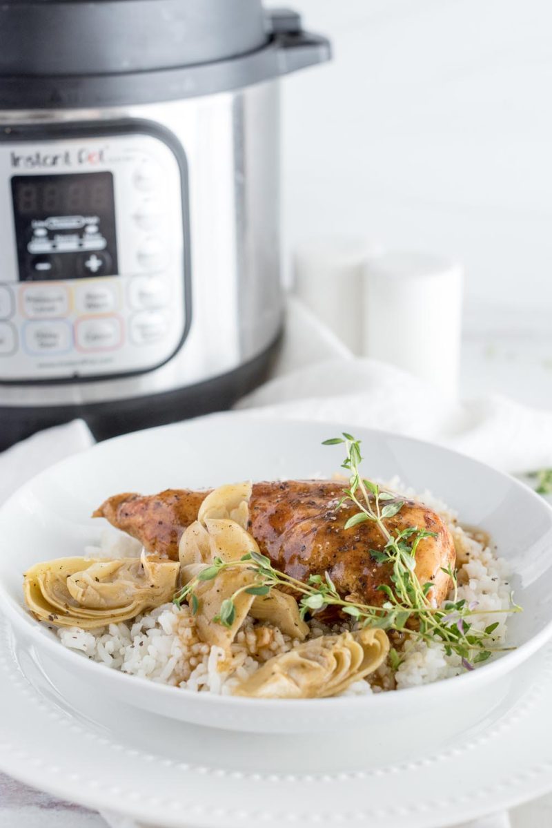 a 45 degree shot with a white bowl filled with artichoke chicken and rice in the foreground, with an Instant Pot visible in the background on the left 