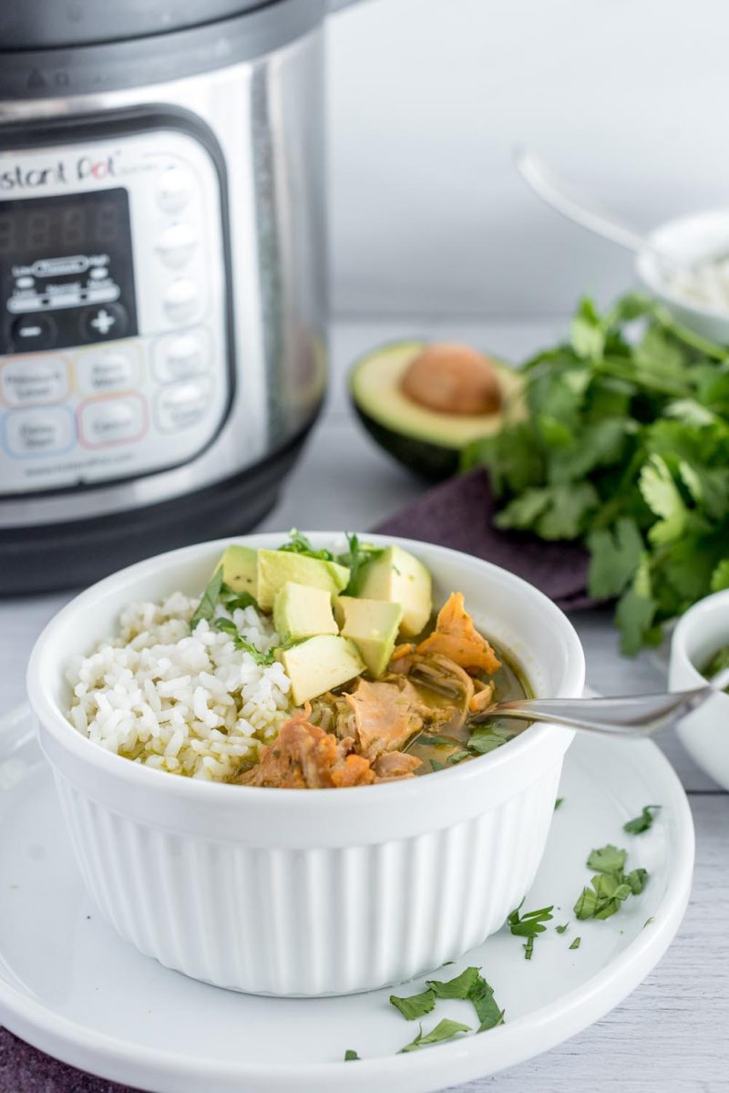 A white bowl filled with asopoa, white rice, and avocado and placed in front of an Instant Pot with fresh cilantro an an avocado in the background.