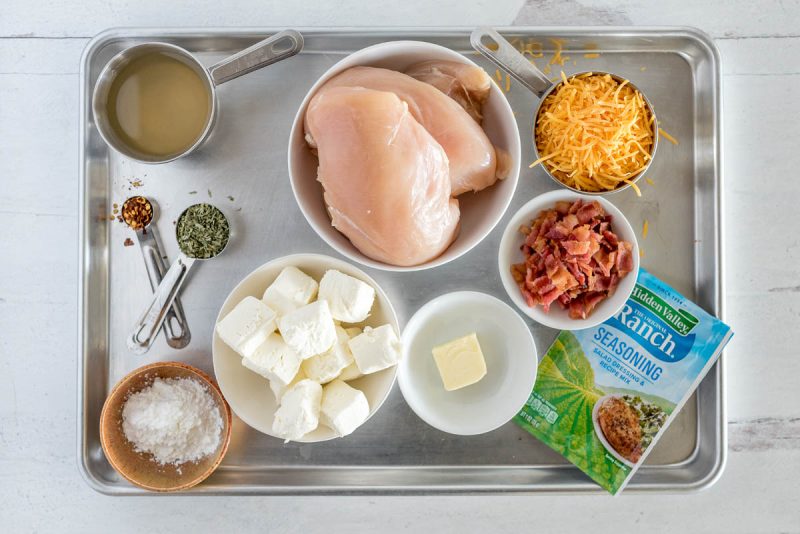 Ingredients for Instant Pot bacon ranch chicken, including ranch seasoning, butter, cream cheese, corn starch, seasonings, chicken broth, chicken breasts, bacon, and shredded cheese.