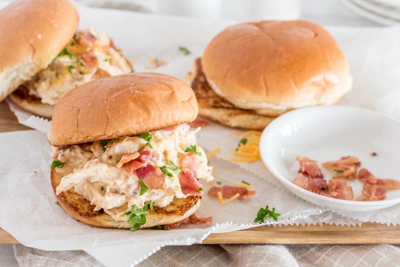 Instant Pot chicken ranch sandwiches served on a bun with bacon, and a bowl of bacon crumbles in the background.