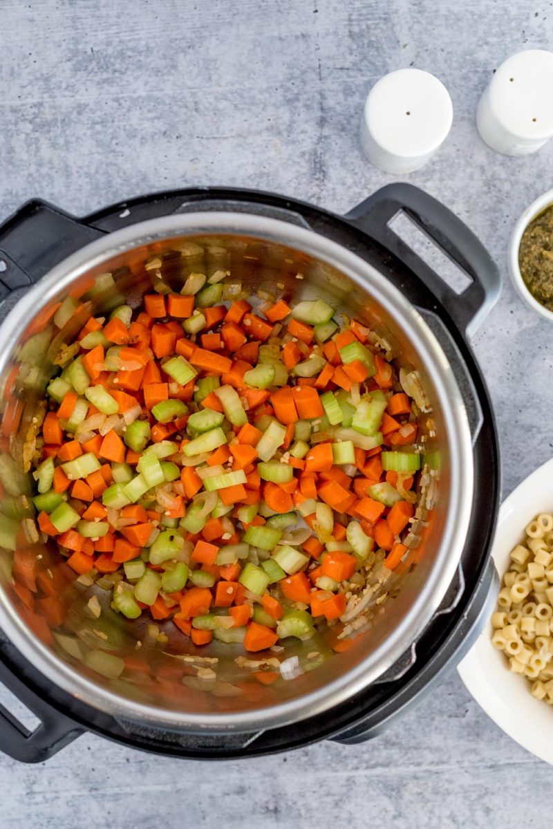 An overhead shot of carrots, onions, and celery being softened in an Instant Pot. A small white shaker of salt and pepper are visible above the Instant Pot, and a small bowl of ditalini is partially in frame on the lower right corner of the photo.
