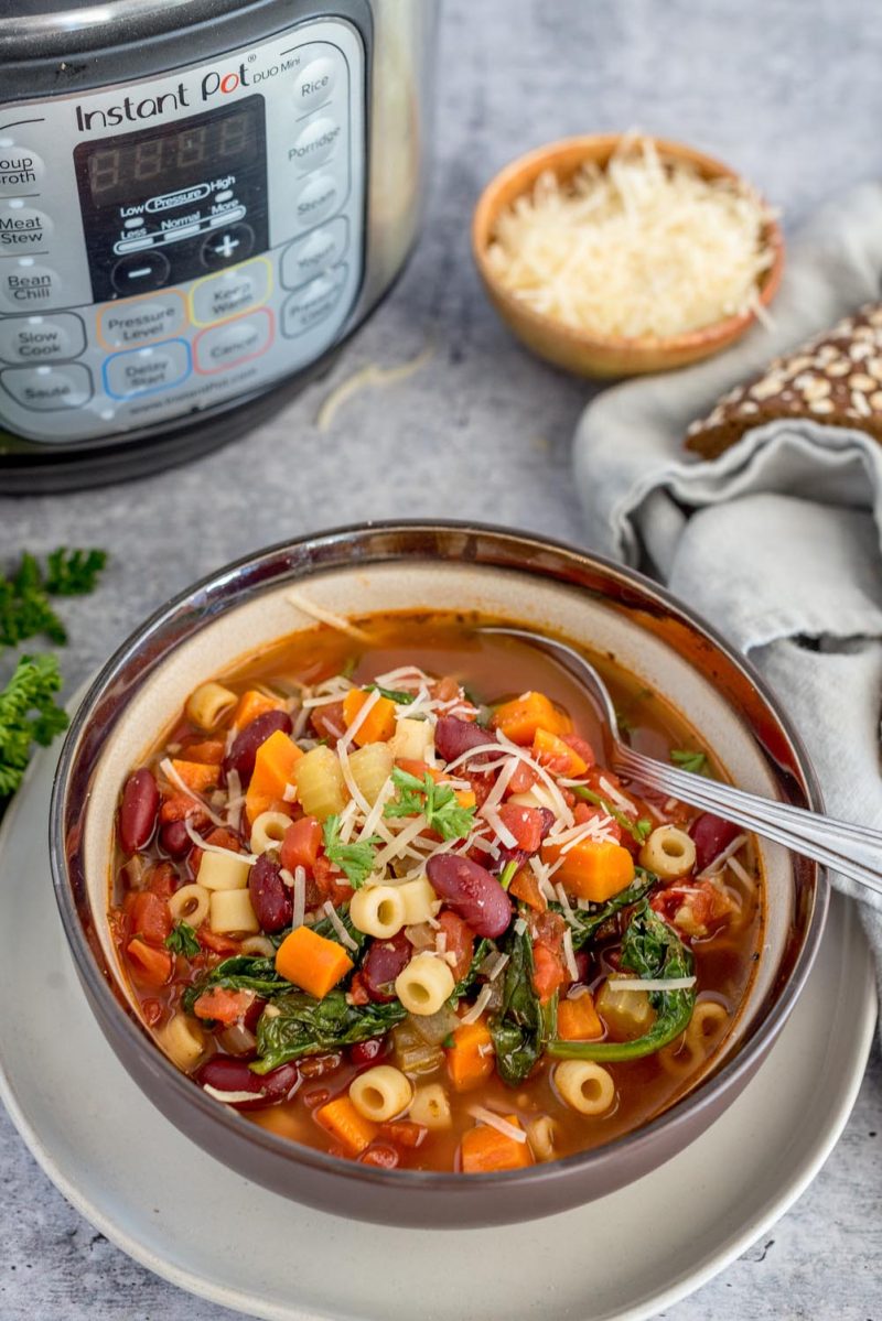 An overhead shot of a bowl of minestrone soup, with a small Instant Pot visible in the background, along with some fresh parsley, a small bowl of grated cheese, and a small bread basket filled with a gray napkin and dark brown slices of whole-grain bread. 