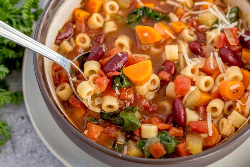 A close-up shot of minestrone soup that really showcases the texture of the dish. A sliver spoonful of soup is along the left-center of the photo, and the ditalini, wilted spinach, red tomatoes and kidney beans, and orange carrots are visible throughout the bowl