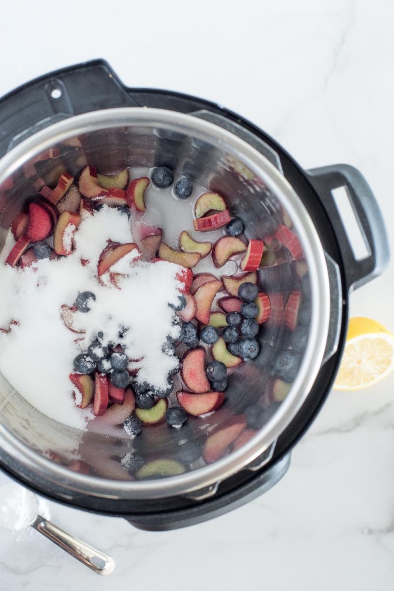 An overhead vertical photo looking into a silver Instant Pot cooking pot. Rhubarb is sliced into crescents and whole blueberries are in the bottom of the pot, along with sugar and lemon juice. A cut lemon is at the bottom left and a tablespoon cornstarch is at the bottom right.