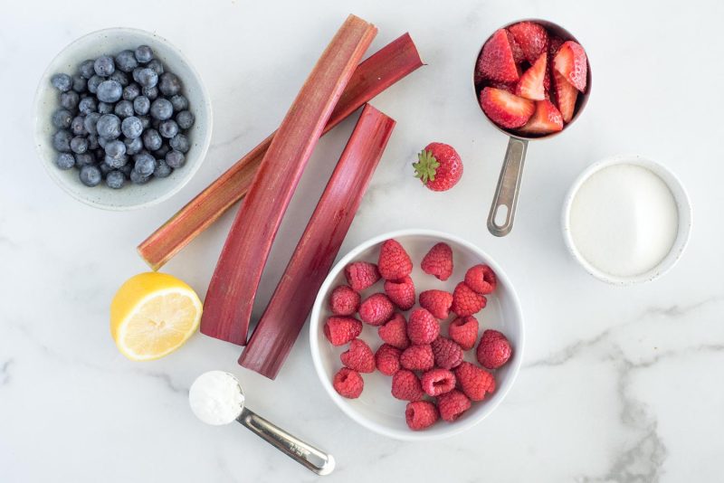 An overhead shot of the ingredients needed to make Berry Rhubarb Sauce in the Instant Pot set out on a white marble background. From left to right, a small bowl of whole fresh blueberries, above a sliced lemon and a tablespoon of cornstarch; three rhubarb stalks; a wide shallow bowl of raspberries; a single whole strawberry with green leaves still on; an old silver measuring cup with hulled and quartered strawberries; and a small, white bowl of granulated sugar.
