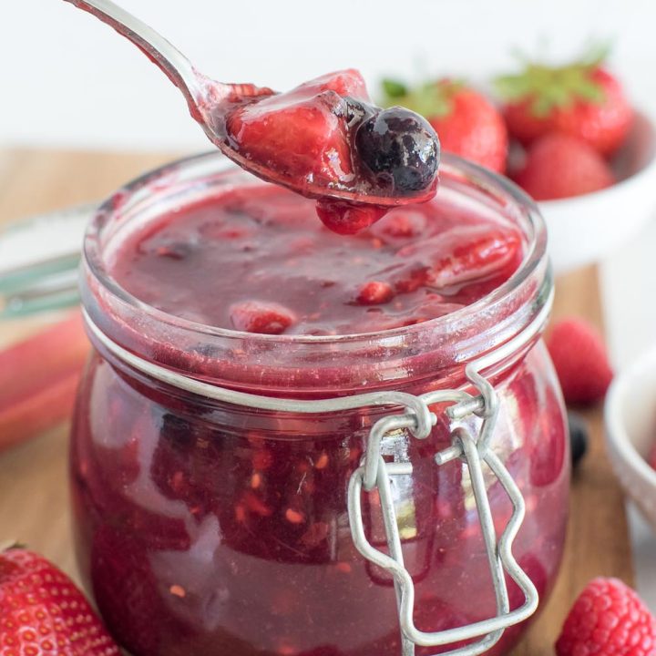 A close-up, shallow-focus vertical shot of a silver spoon filled with strawberries and blueberries suspended over a jar of deep red Berry Rhubarb Sauce. Fresh fruit garnishes the wooden cutting board at the very bottom of the picture.