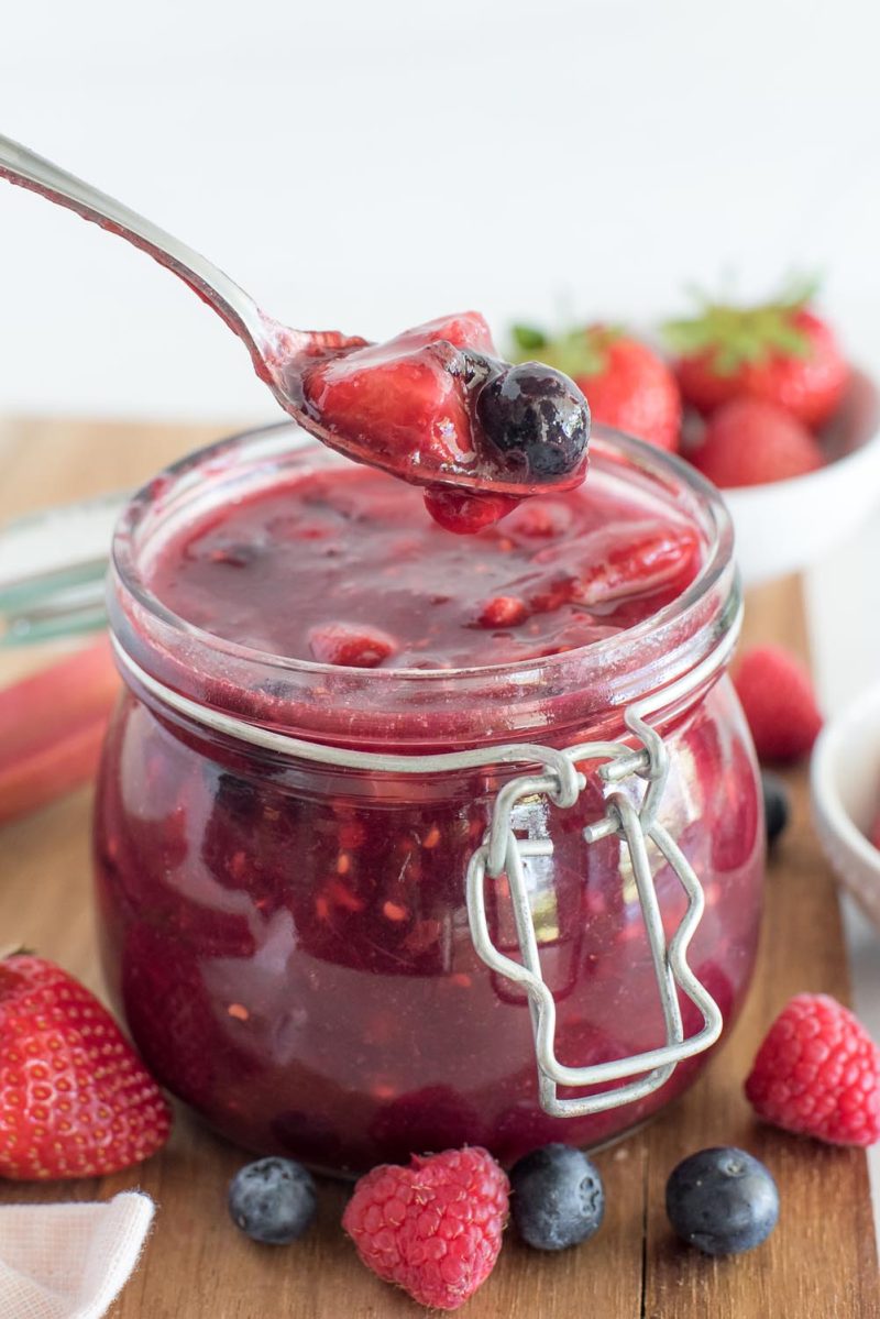 A close-up, shallow-focus vertical shot of a silver spoon filled with strawberries and blueberries suspended over a jar of deep red Berry Rhubarb Sauce. Fresh fruit garnishes the wooden cutting board at the very bottom of the picture.
