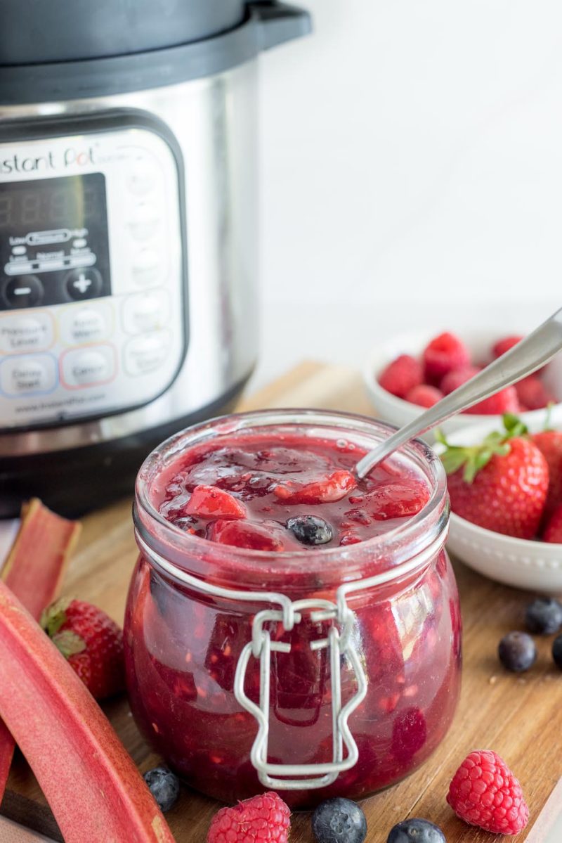 A 45-degree vertical shot with a jar filled with a lovely red berry rhubarb sauce. Fresh rhubarb stalks are in the lower-left, with fresh raspberries, blueberries, and strawberries along the bottom and right edge. An Instant Pot is slightly out of focus in the background.