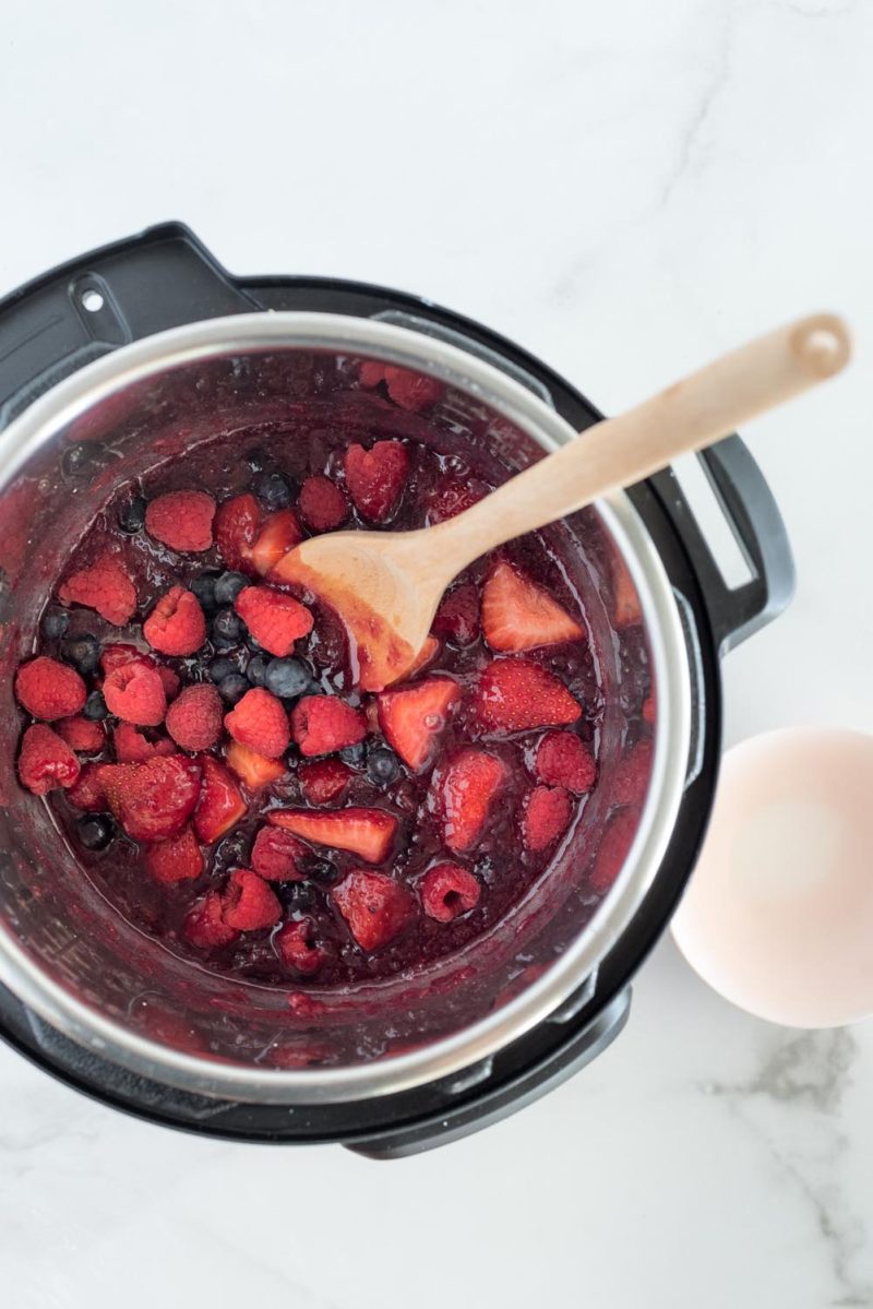An overhead shot looking into the Instant Pot filled with the rhubarb compote and the fresh raspberries, strawberries, and blueberries added. A wooden spoon rests against the pot edge on the upper right.
