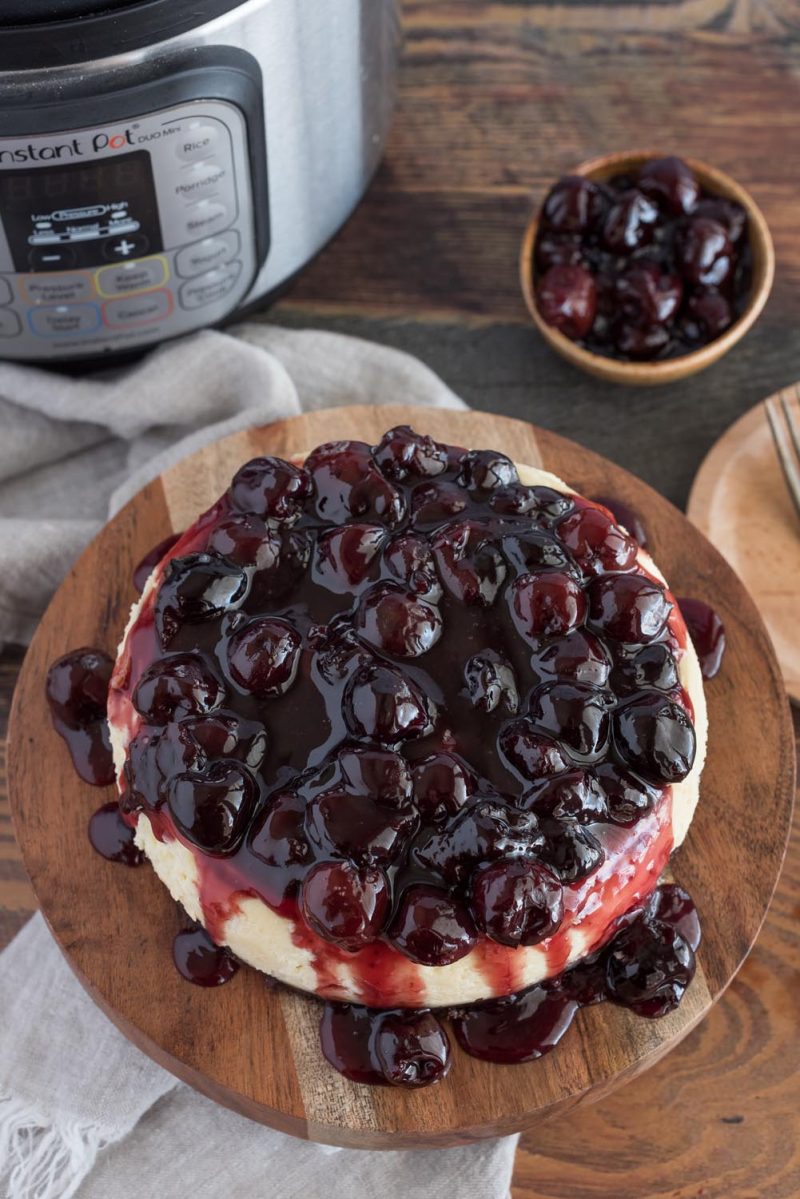 Overhead picture of a black forest cheesecake on a wooden serving plate placed in front of an Instant Pot.