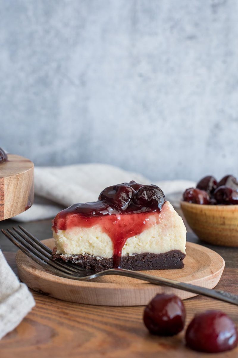 A side shot of a slice of Cherry Cheesecake, made with a brownie crust, vanilla cheesecake, and topped with cherry compote. Sitting on a wooden round with a dark stainless fork, with a bowl of cherries and extra cheesecake in the background.