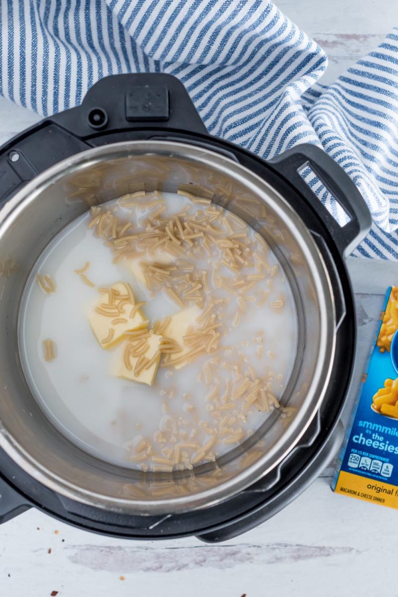 An overhead shot of an Instant Pot cooking pot filled with butter, milk, water, and pasta. A blue and white striped towel and a box of mac and cheese are visible in the background.