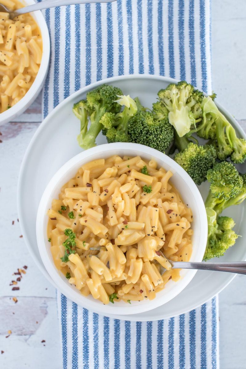 An overhead vertical shot of a white bowl of macaroni and cheese sitting on a white plate filled with chopped broccoli. The plate is sitting on a blue and white striped hand towel.