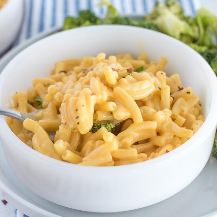 A close-up horizontal photo of a white bowl filled with yellow store-bought mac and cheese, garnished with red pepper flakes and parsley. The bowl is resting on a white plate with broccoli on the side.