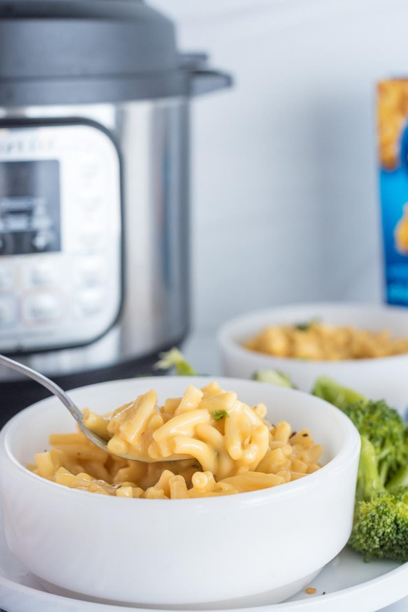 A vertical photo, with a silver spoon coming in from the left side of the frame to scoop a bite of macaroni and cheese from a white bowl. The bowl is resting on a white plate with broccoli on the side. An Instant Pot is out of focus in the background, along with a blue box of Kraft Macaroni and Cheese just visible on the right side of the frame.