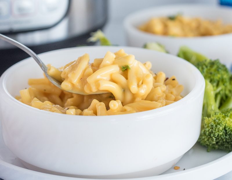A vertical photo, with a silver spoon coming in from the left side of the frame to scoop a bite of macaroni and cheese from a white bowl. The bowl is resting on a white plate with broccoli on the side.