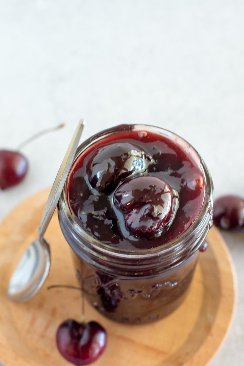 a mason jar filled with Instant Pot cherry topping sitting on a wooden plate with a silver spoon propped along the left side of the jar.