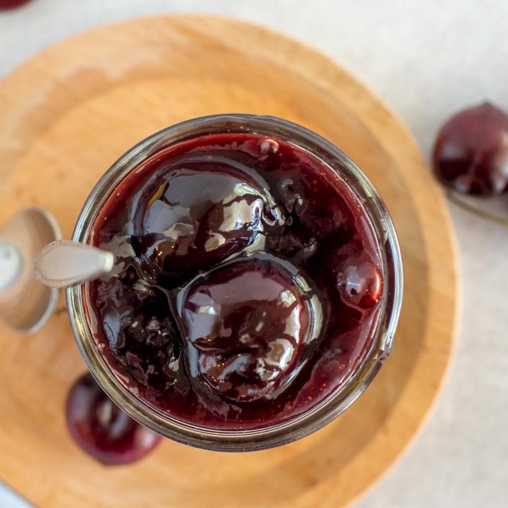 An overhead shot looking into the a jar of cherry sauce, with a wooden background and resh cherries scattered alongside the dish.