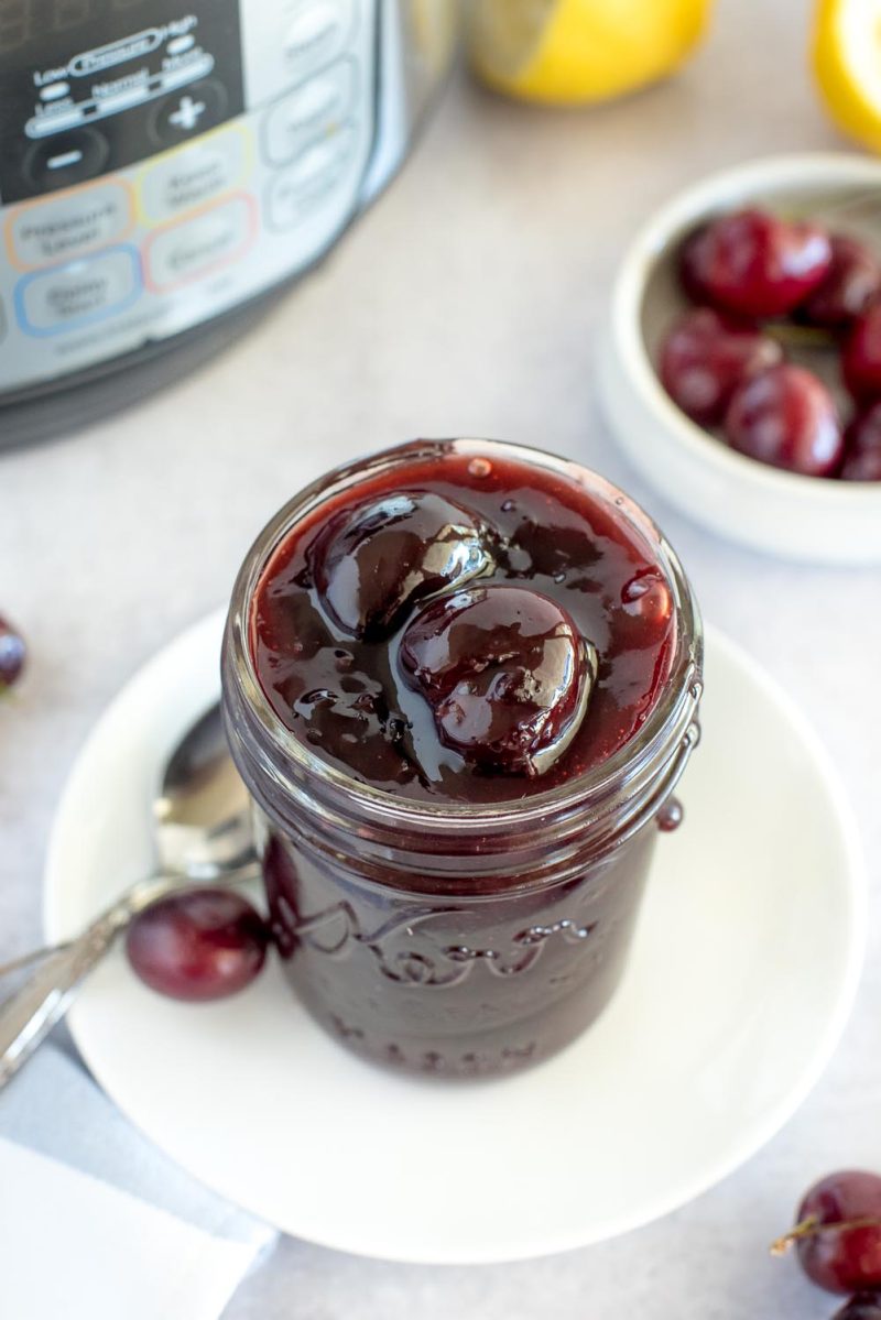 a mason jar sitting on a white plate, filled to the brim with cherry compote. An Instant Pot, lemons, and fresh cherries are out of focus along the top background.