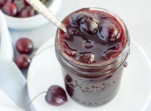 A mason jar filled to slightly overflowing with cherry compote, with a silver spoon ready to scoop.