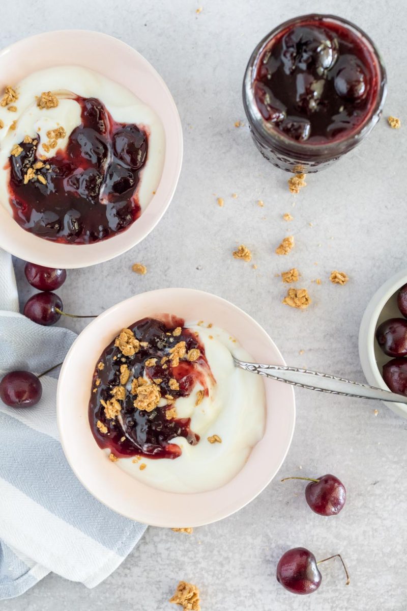 An overhead shot of two pale pink bowls of plain yogurt topped with cherry compote and sprinkled with granola. With extra granola and cherries scattered on the background.