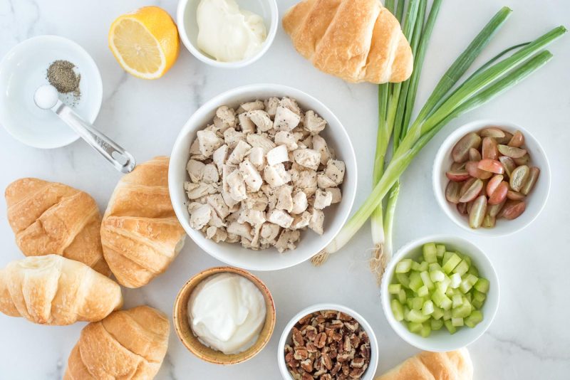 Ingredients for Instant Pot chicken salad sandwiches including croissants, mayonnaise, pecans, celery, grapes, green onions, salt, pepper, lemons, and diced chicken.