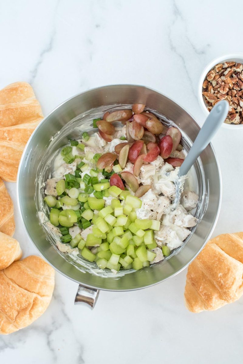 Mixing the chicken salad in a metal bowl with celery, green onion, grapes, chicken and mayonnaise, with pecans and croissants in the background.