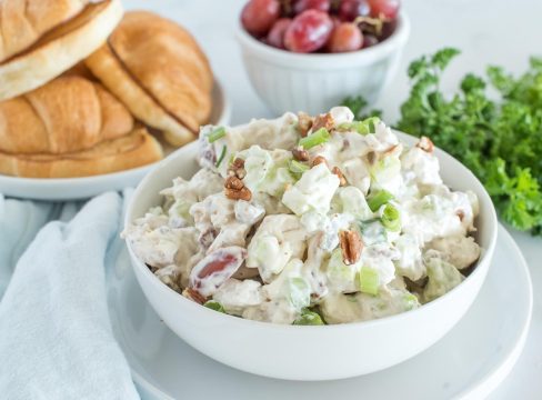 picture of a bowl of chicken salad mix, with a bowl of grapes, croissants, and fresh parsley in the background.