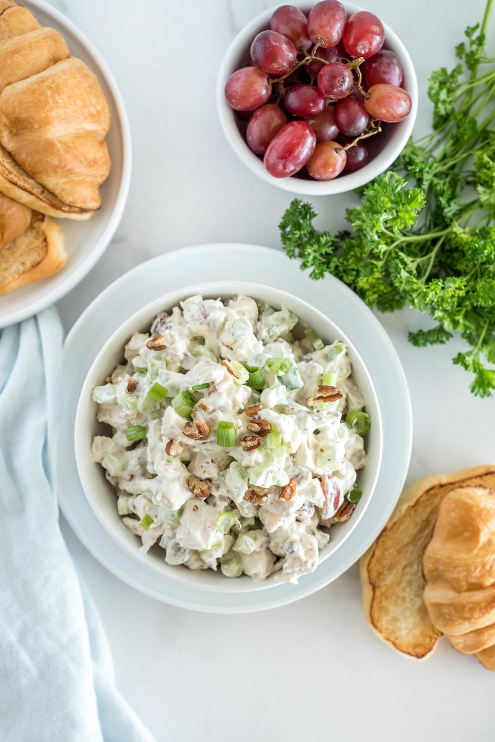 Overhead picture of a bowl of chicken salad mix, with a bowl of grapes, croissants, and fresh parsley in the background.