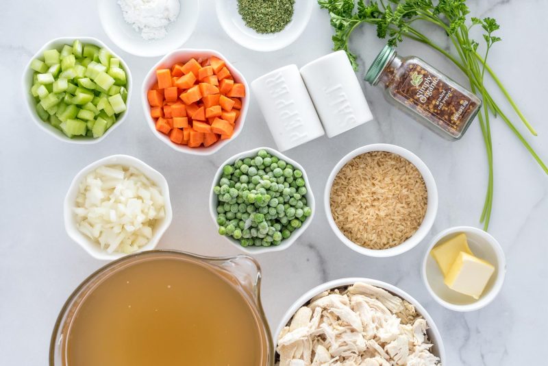 an overhead shot of the ingredients needed to make chicken and rice soup, including chicken broth, shredded rotisserie chicken, butter, rice, peas, carrots, celery, onions, spices, and parsley.