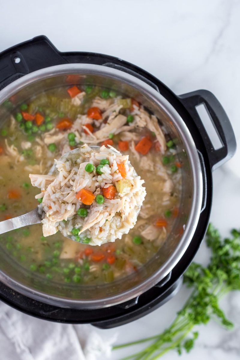 An overhead shot looking down onto a silver ladle heaped with chicken, rice, carrots, peas, suspended over and Instant Pot filled with chicken and rice soup