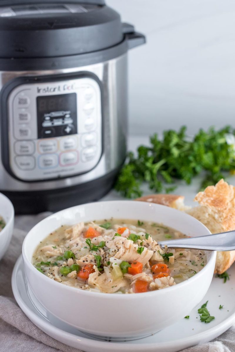An chicken soup in a white bowl filled with rice, carrots, celery, and potatoes, with an Instant Pot, parsley, and a torn roll in the background.