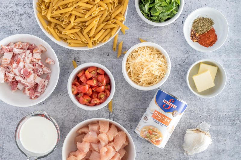 Ingredients for Instant Pot chicken bacon penne, including diced chicken, chicken broth, garlic, heavy cream, butter, parmesan cheese, diced tomatoes, diced bacon, spices, chopped spinach, and penne pasta.