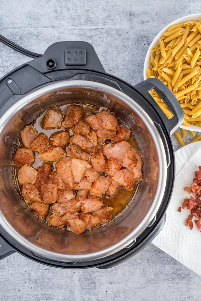 Meat, spices, and broth in the bottom of the Instant Pot for making chicken bacon penne.