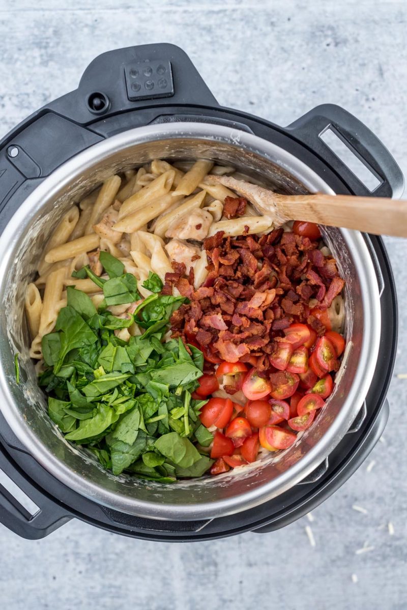 An overhead picture of chicken bacon penne cooked in an Instant Pot stirring in the bacon, tomato, and chopped spinach.