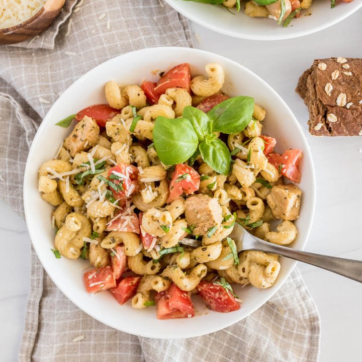 Over head shot of a white bowl with Instant Pot chicken pesto pasta topped with fresh basil and parmesan cheese, with bread and more basil in the background.