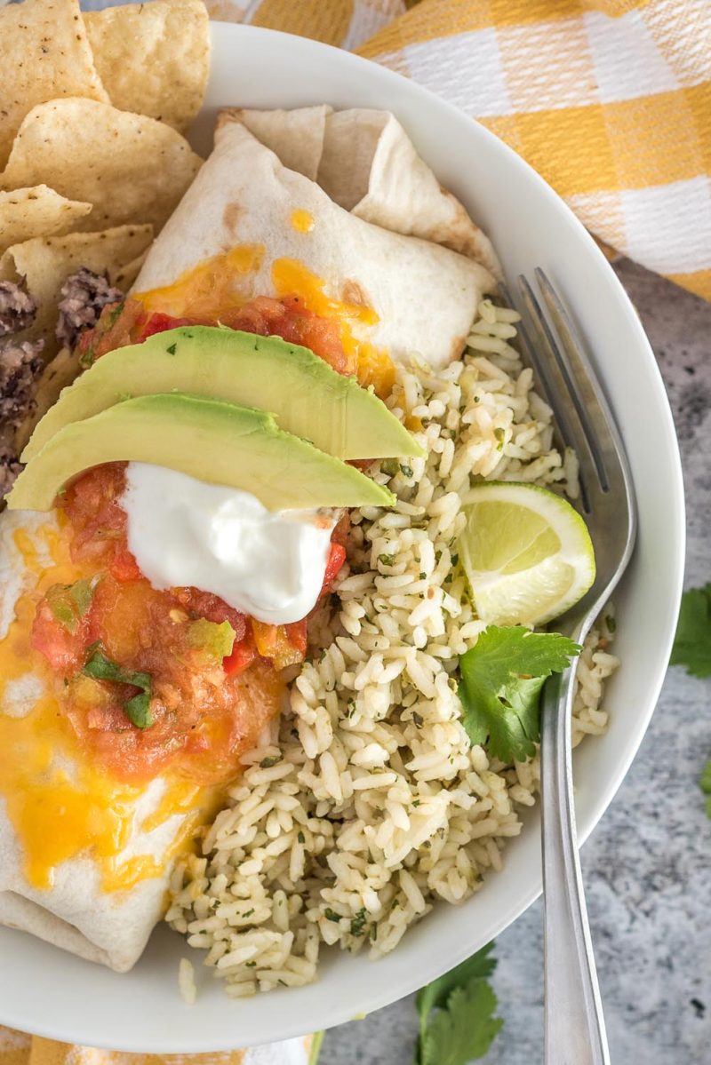 a close-up vertical overhead photo of a shallow white bowl filled with a wrapped chicken tinga enchilada covered with melted cheese, tomato salsa, sour cream, and avocado garnish, with a lime wedge and a cilantro garnish on green rice.