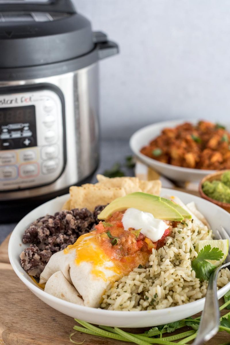 A 45 degree photo of chicken tinga with black beans and rice in front of an Instant Pot on the background left and a serving bowl of chicken tinga enchilada filling on the right.