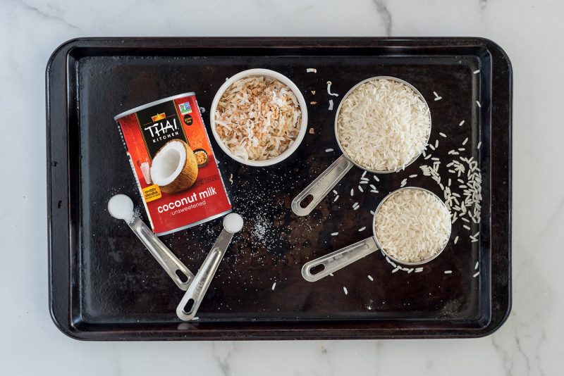 An overhead shot of a dark baking sheet with the ingredients for making coconut rice displayed. From left to right, a teaspoon of sugar, a can of coconut milk, a half teaspoon salt, a white bowl filled with toasted coconut, and one and a half cups of white jasmine rice.