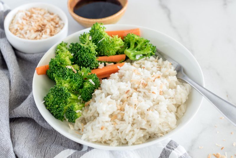 A 45 degree shot of a small white bowl filled with coconut rice garnished with toasted coconut, with broccoli and carrots along side, with a fork tucked into the rice on the right. In the top of the photo are a small white bowl of toasted coconut, and a small tan bowl of dark soy sauce.