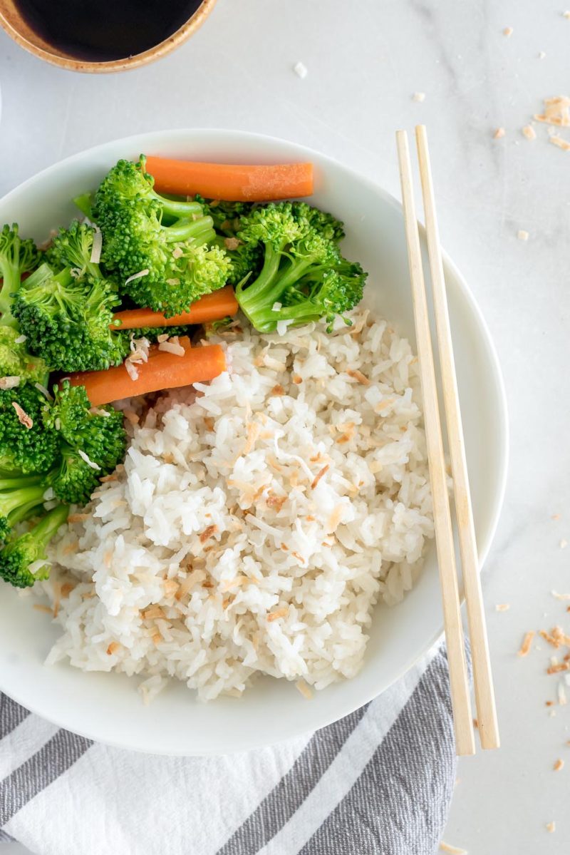 An overhead shot of prepared coconut rice, offset slightly so the left side of the bowl, filled with broccoli and carrots, is cut out of the frame. The focus is on the rice, with a pair of chopsticks set side by side on the right side of the bowl.