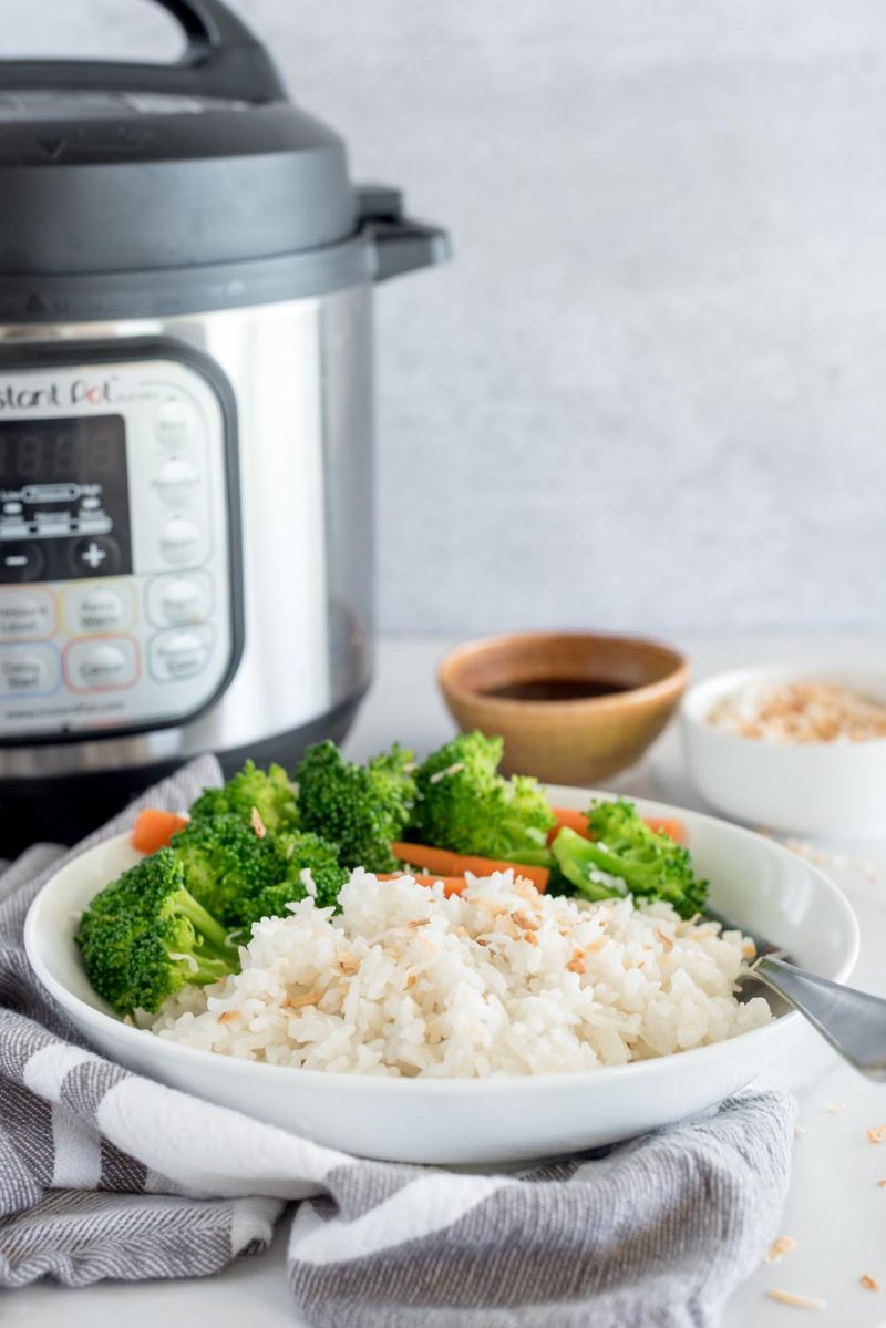 A front shot of a bowl of coconut rice set on a gray napkin in front of an Instant Pot, with an out of focus bowl of soy sauce and a white bowl of toasted coconut set to the right side of the Instant Pot.