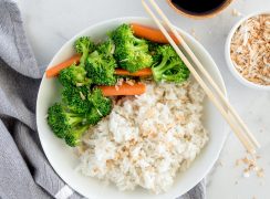 An overhead shot of a small white bowl filled with coconut rice garnished with toasted coconut, with broccoli and carrots along side, with a set of chopsticks at an angle on the top right side of the bowl. The bottom of the white bowl is slightly cut out of the bottom of the frame.