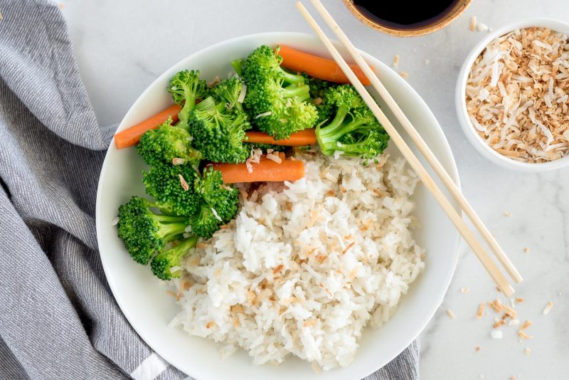 An overhead shot of a small white bowl filled with coconut rice garnished with toasted coconut, with broccoli and carrots along side, with a set of chopsticks at an angle on the top right side of the bowl. The bottom of the white bowl is slightly cut out of the bottom of the frame.