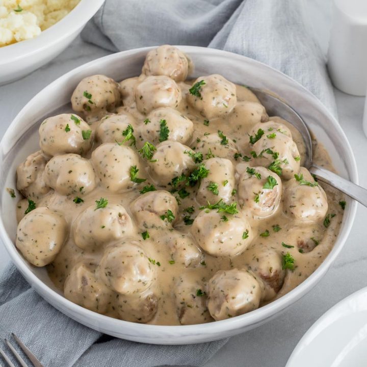 Instant Pot Swedish Meatballs and gravy in a serving bowl, and mashed potatoes in the background.