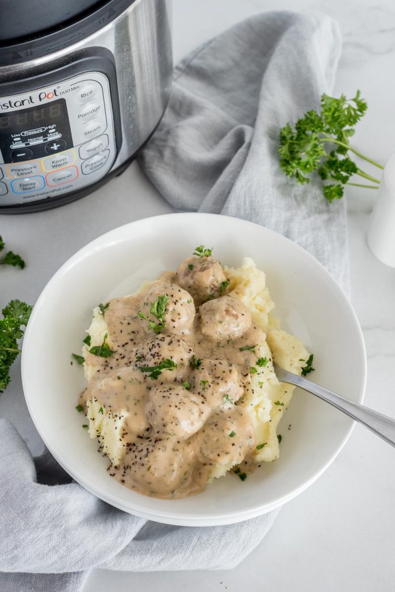 A bowl of mashed potatoes topped with Swedish Meatballs in a white bowl, placed in front of an Instant Pot.